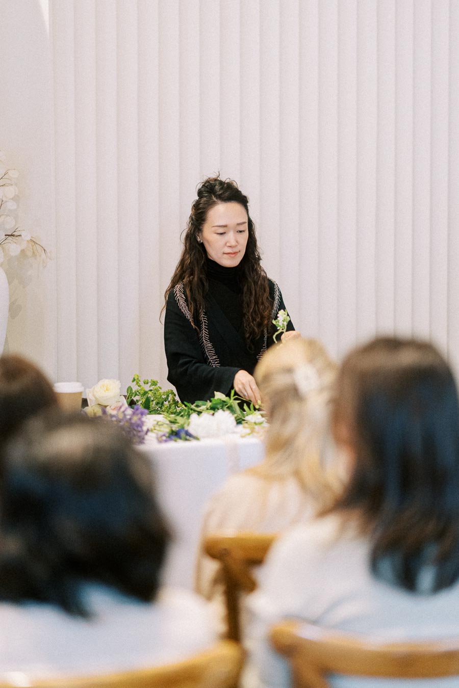 A woman demonstrating floral arrangement techniques at a workshop, with attendees watching closely and flowers on the table.