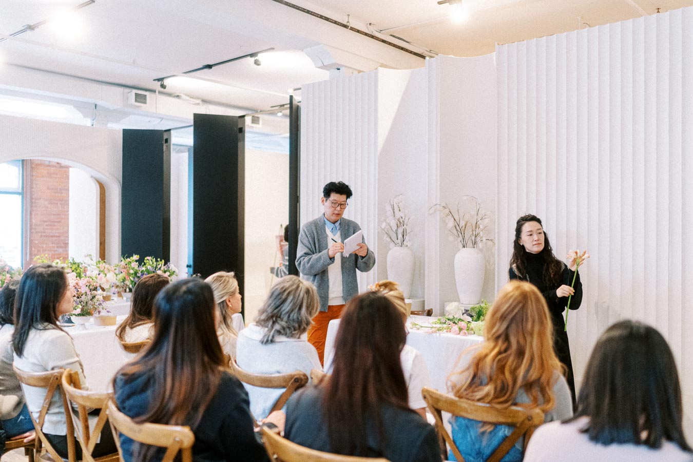 Workshop setting with a diverse group of people seated, attentively watching a floral arrangement demonstration by two presenters in a bright room.