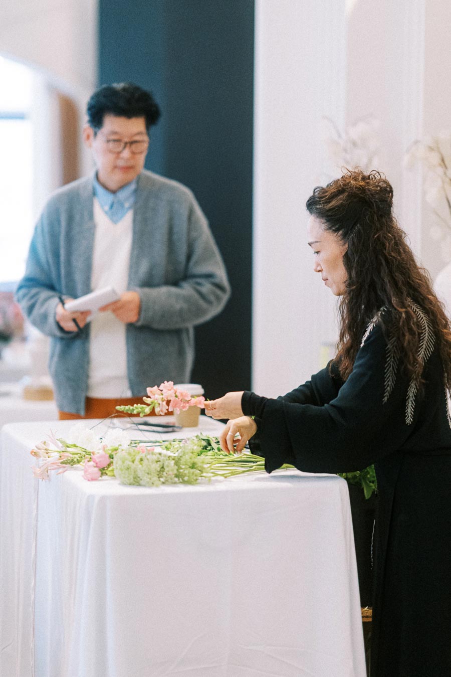 A woman arranging a floral bouquet at a table, with a man in the background taking notes, demonstrating a flower workshop setting.