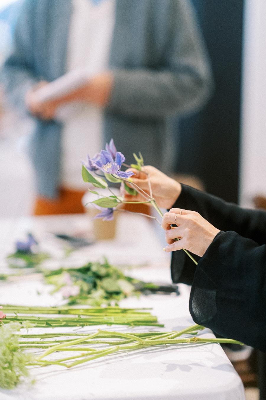 A person arranging delicate purple flowers on a table, with scattered green stems and blurred background, showcasing a floral workshop or event.