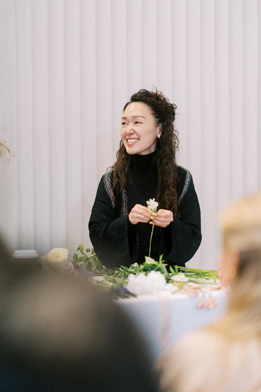 A woman smiling while arranging flowers at a table, wearing a black sweater, in a bright indoor setting. Fresh flowers and vibrant greenery are spread out before her, creating a serene and creative atmosphere.