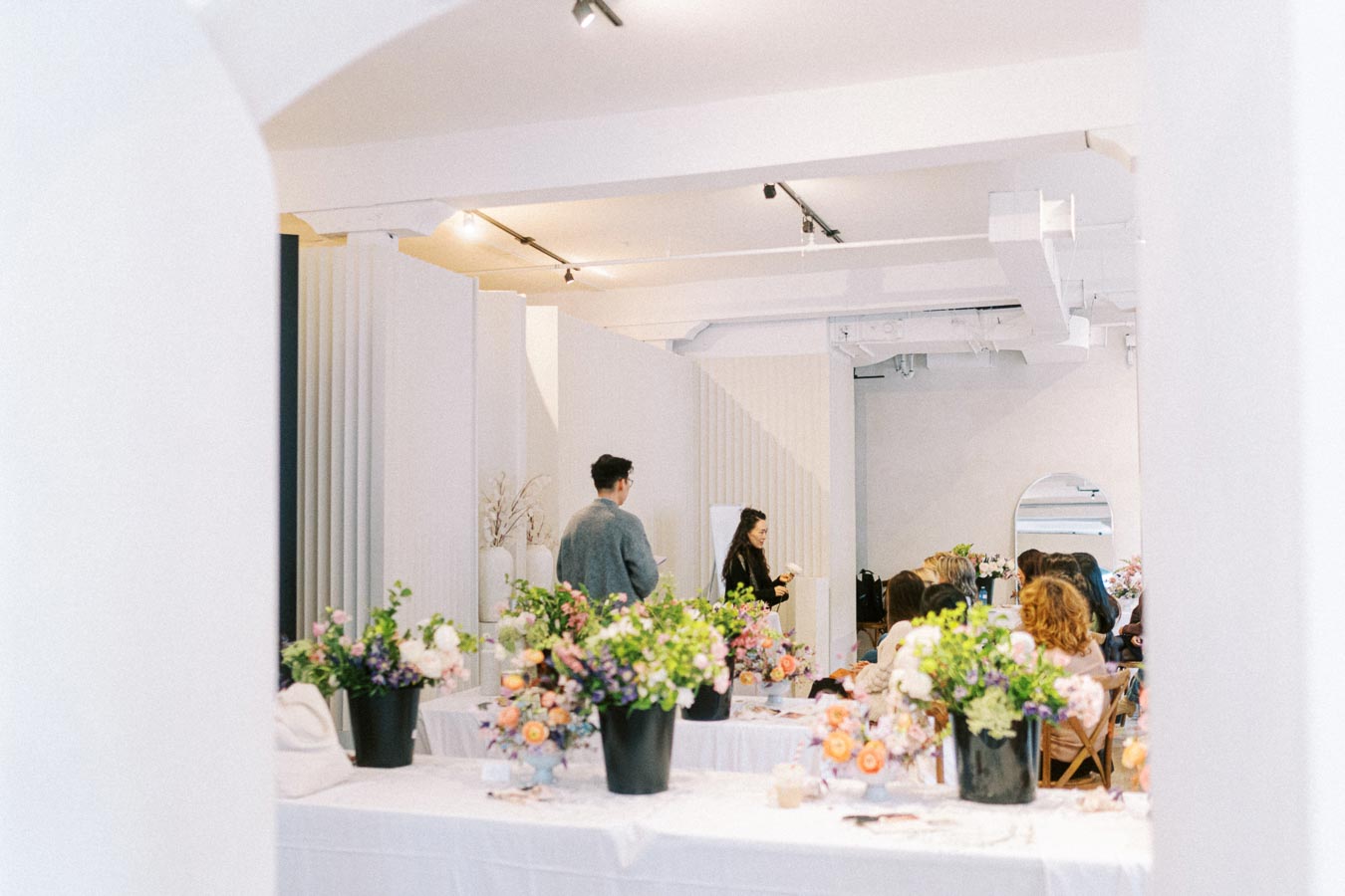 A bright, minimalistic event space with tables adorned with vibrant flower arrangements. A speaker addresses a small audience, emphasizing a creative and intimate atmosphere.