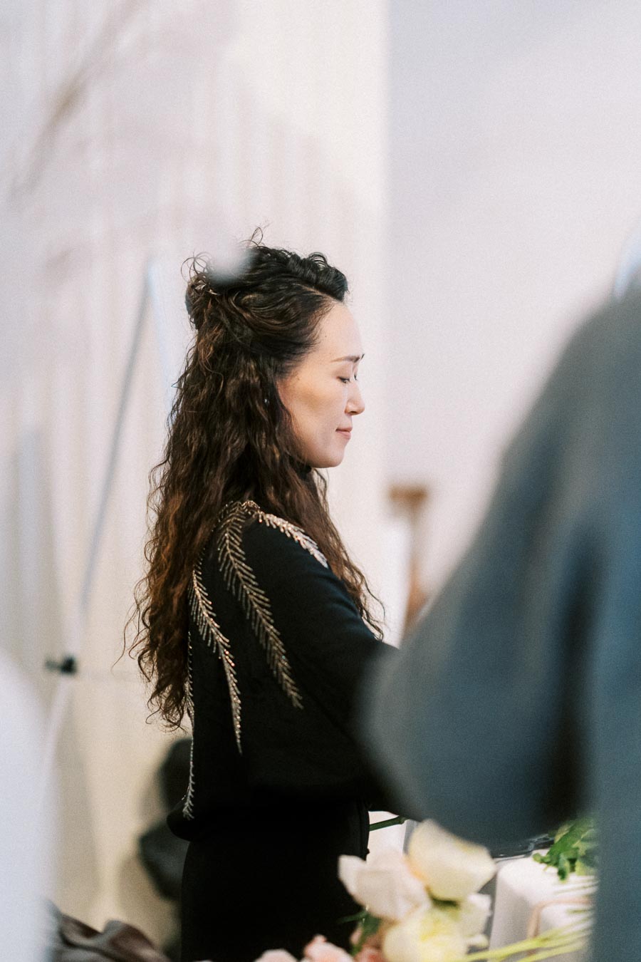 A woman with long curly hair stands indoors with eyes closed, wearing a black top with intricate feather-like embroidery. Soft natural light and flowers are visible in the foreground, creating a serene atmosphere.