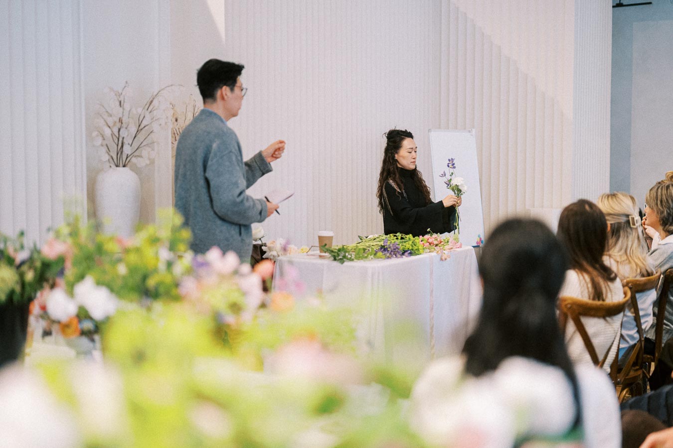 Workshop setting with a woman teaching floral arrangement to an attentive audience, featuring colorful flowers and plants on display.