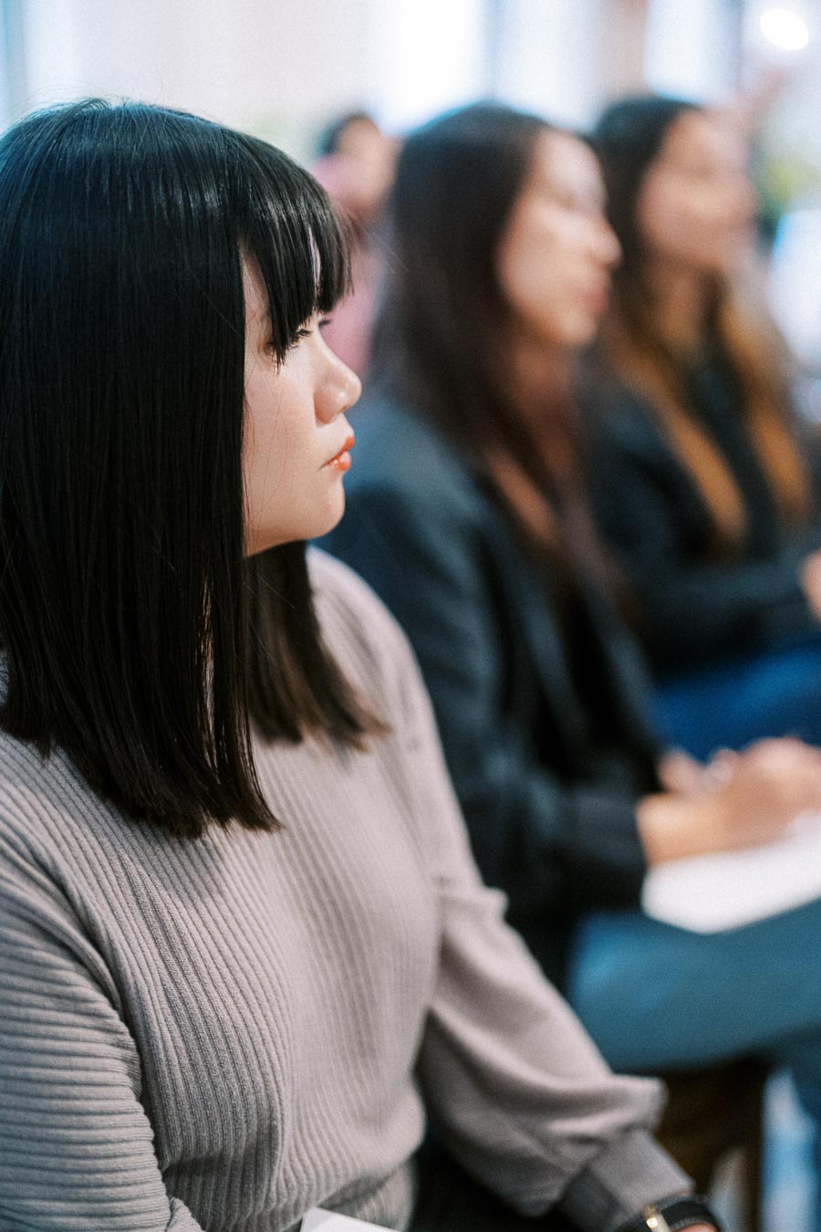 A focused woman attentively listens during a seminar or conference, wearing a gray sweater, with others in the background in a blurred setting.