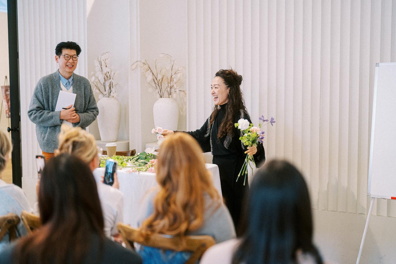 A woman conducting a floral arrangement workshop, holding a bouquet, while a man stands beside her with a notepad. An attentive audience observes and takes notes in a modern, minimalist setting.