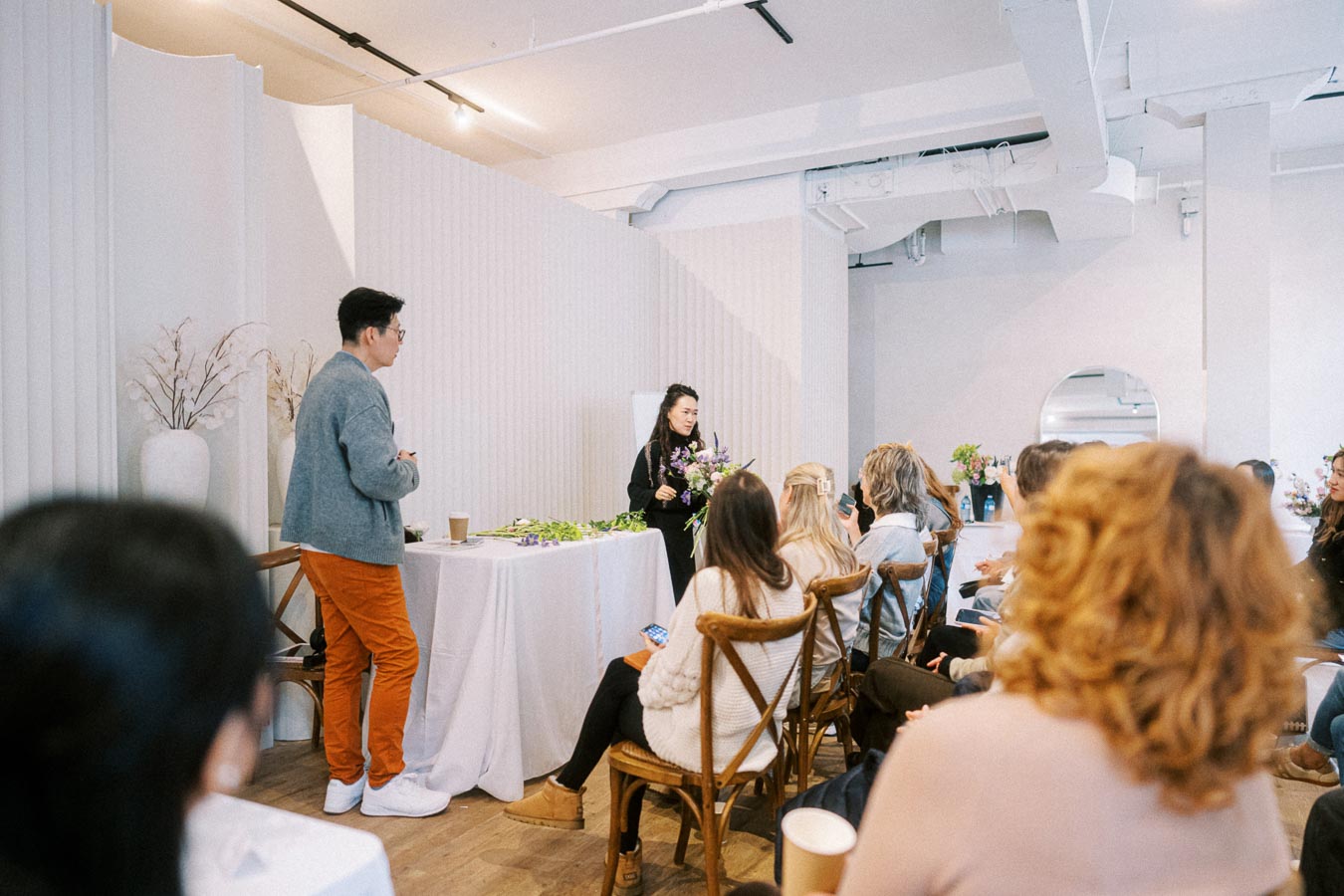 Workshop setting with a female speaker demonstrating floral arrangements to an engaged audience in a modern, bright room.