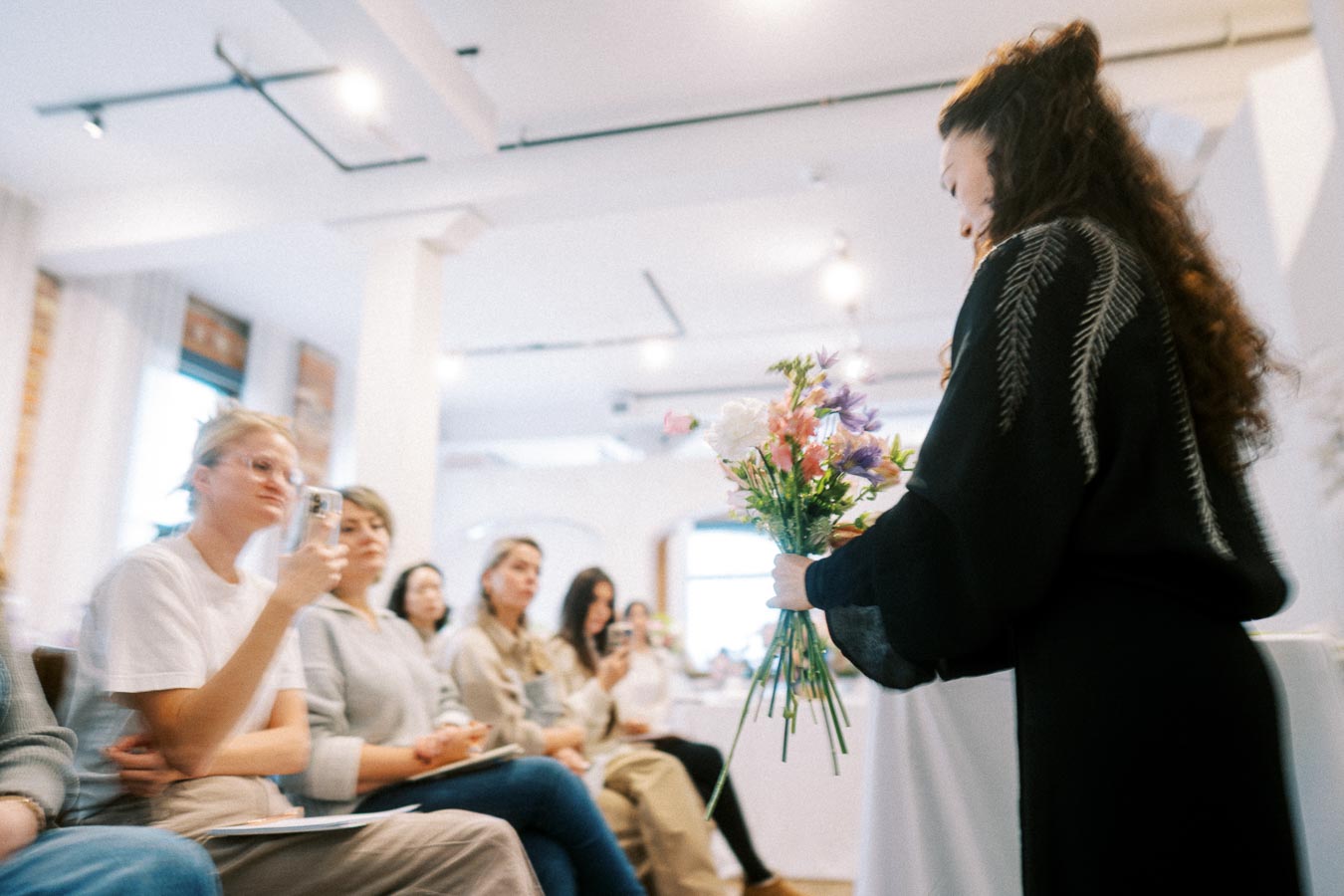 A woman in a dark outfit holds a bouquet of colorful flowers while presenting to an attentive audience in a bright, modern classroom or workshop setting.