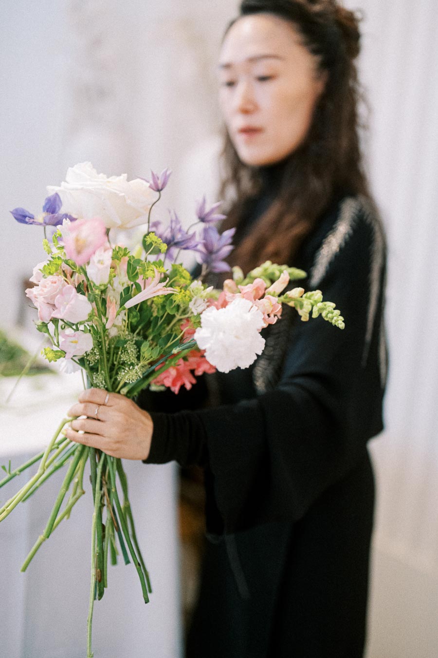 A person holding a vibrant bouquet of assorted flowers, including white roses, purple blooms, and pink carnations, in a softly lit setting.