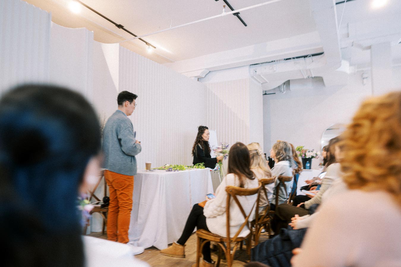 Audience attending a floral arrangement workshop, with an instructor demonstrating techniques at the front. The setting is a bright, modern room with attendees seated attentively.