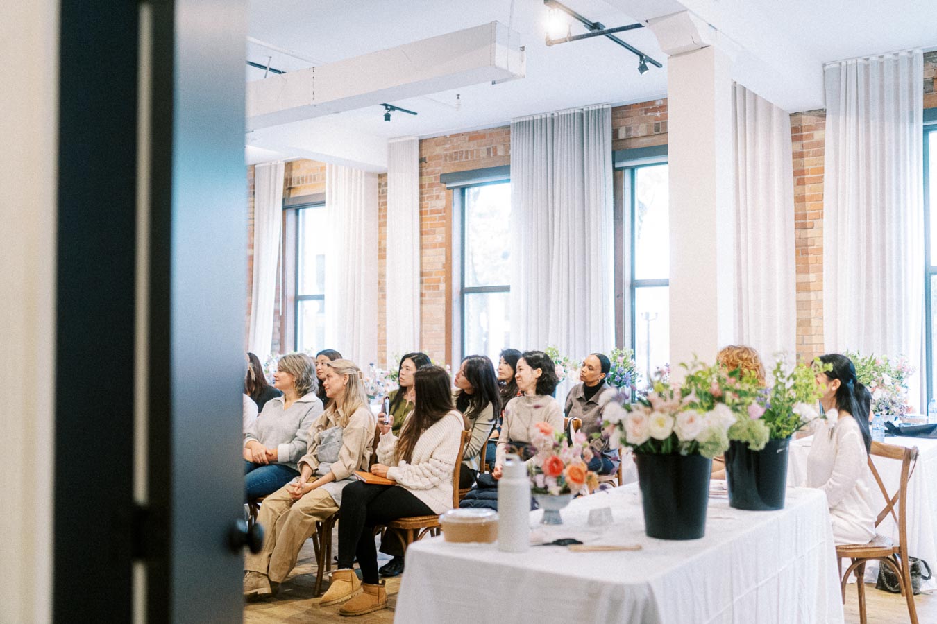 A diverse group of people attentively listening at an indoor workshop, surrounded by bright natural light and floral decorations on tables.