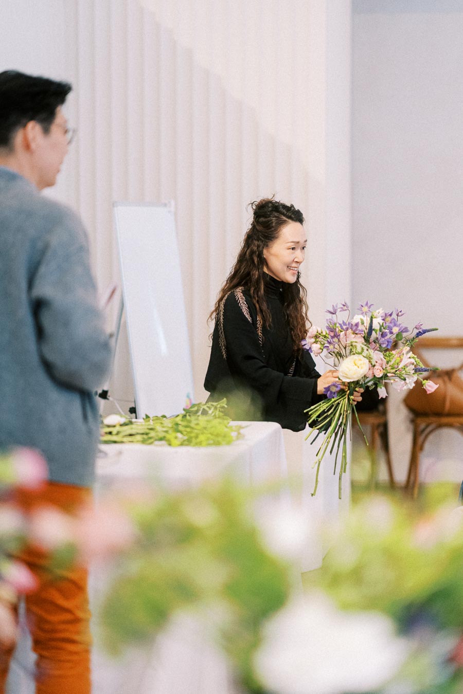 A woman holding a colorful bouquet of flowers during a floral arrangement workshop, with a blurred background of participants and floral materials on a table.