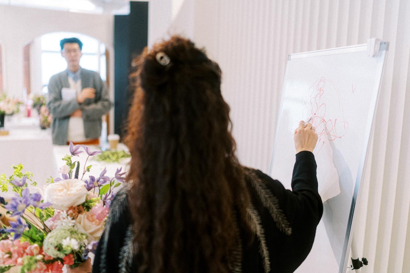 Person with long hair drawing on a whiteboard during a presentation, with colorful flowers on the table in the foreground and a blurred audience member in the background, creating a creative and professional meeting atmosphere.