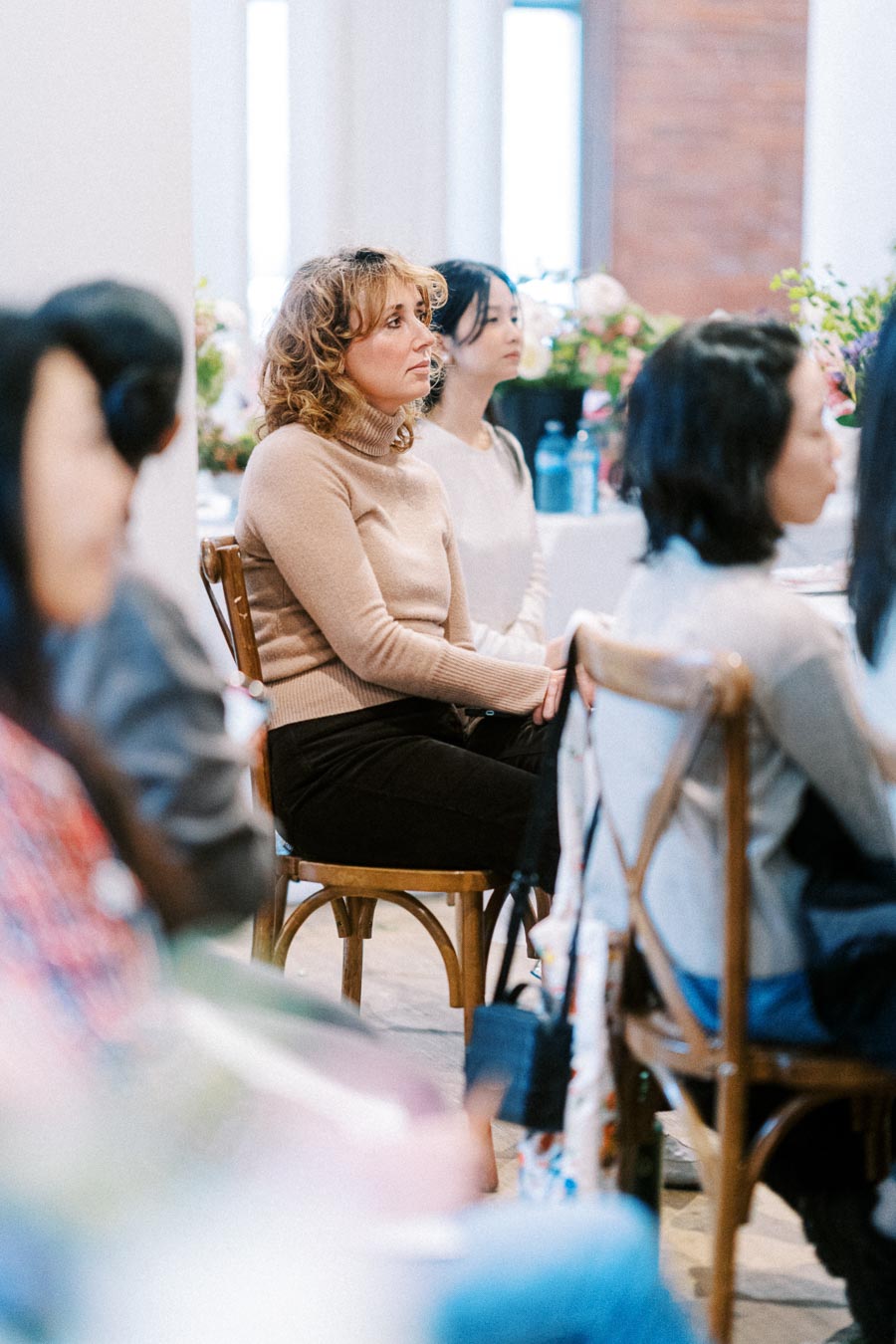 A group of people attentively listening to a speaker at a conference, with a focus on a woman in a beige sweater.
