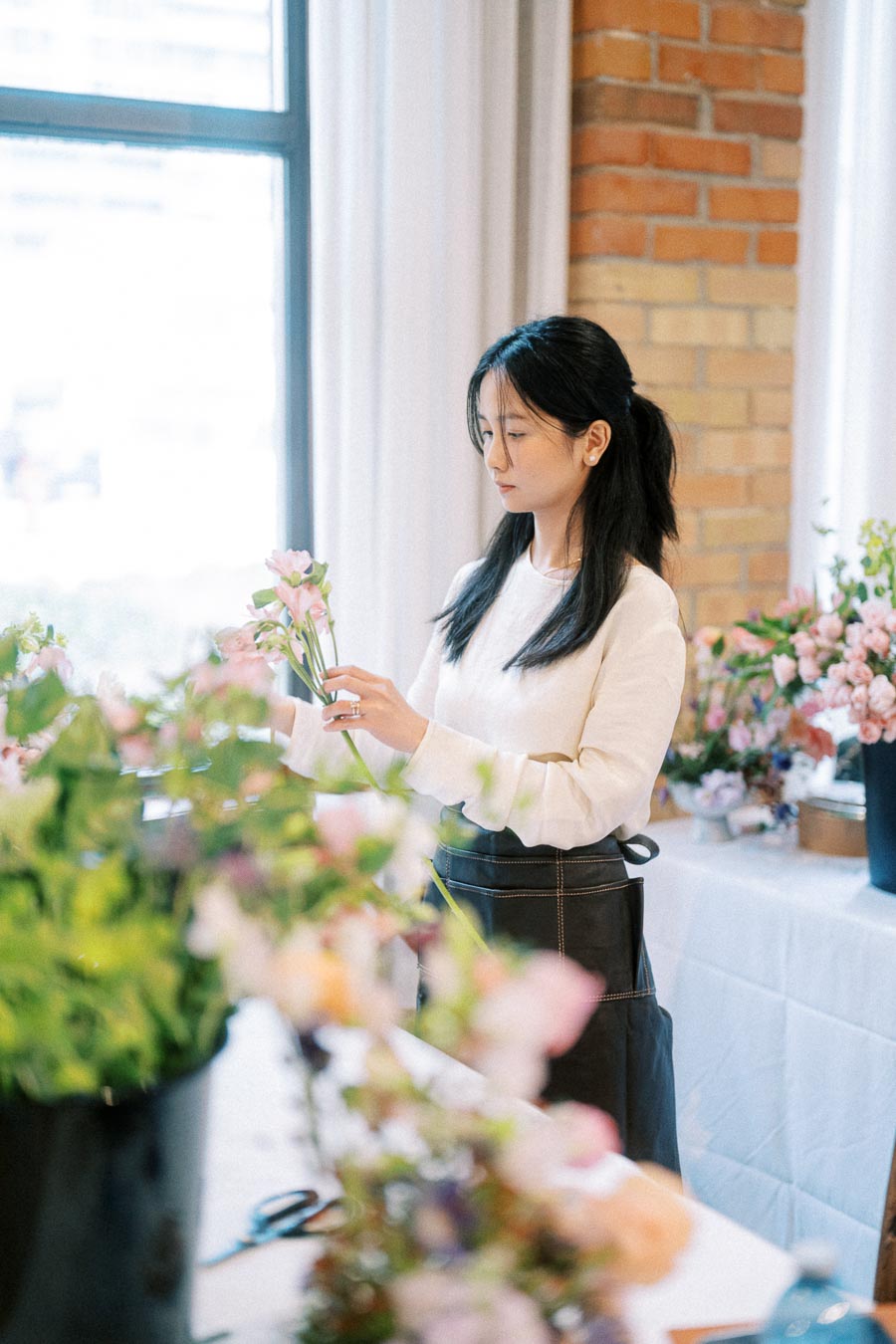 A woman arranging flowers at a workshop, surrounded by colorful blooms, wearing a white shirt and dark apron inside a studio with a brick wall backdrop.