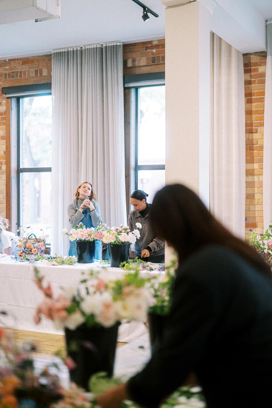 Florists arranging colorful flower bouquets on a table in a bright, airy room with large windows and exposed brick walls.