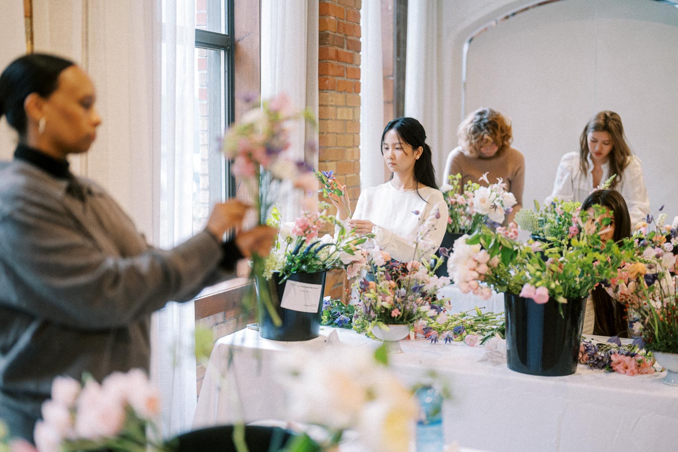 People participating in a floral arrangement workshop, focusing on creating colorful bouquets with various flowers on a long table near large windows in a bright and airy space.