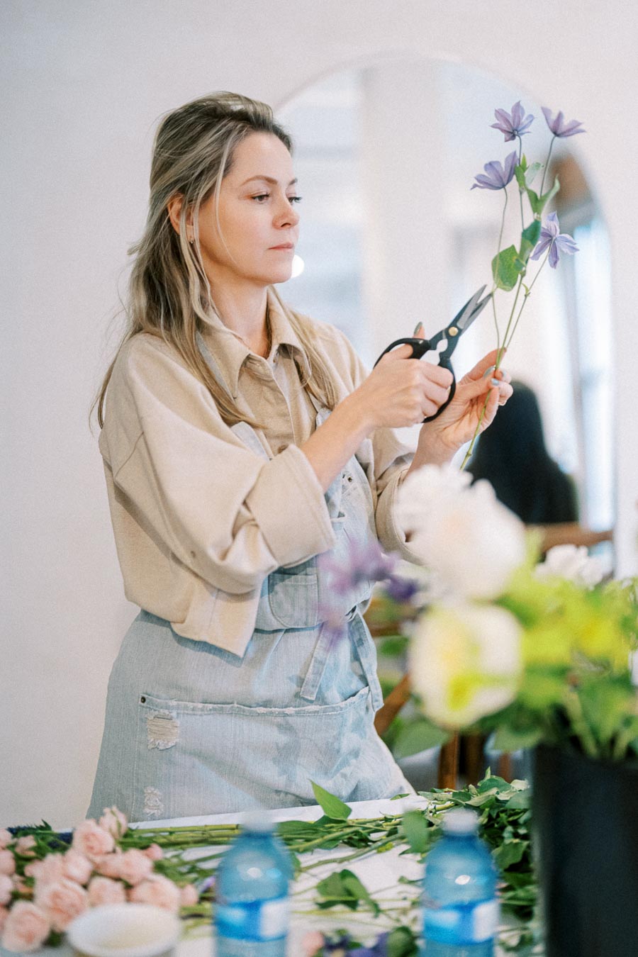 A woman arranging flowers while wearing a beige shirt and blue apron, surrounded by various floral stems and tools on a table.