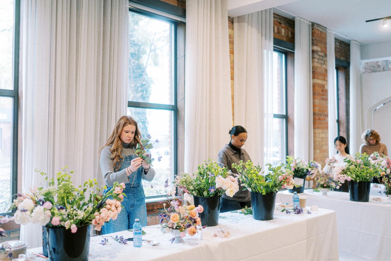 People participating in a flower arrangement workshop, focusing intently on creating floral bouquets with pink, white, and purple flowers in a bright, spacious room with large windows.