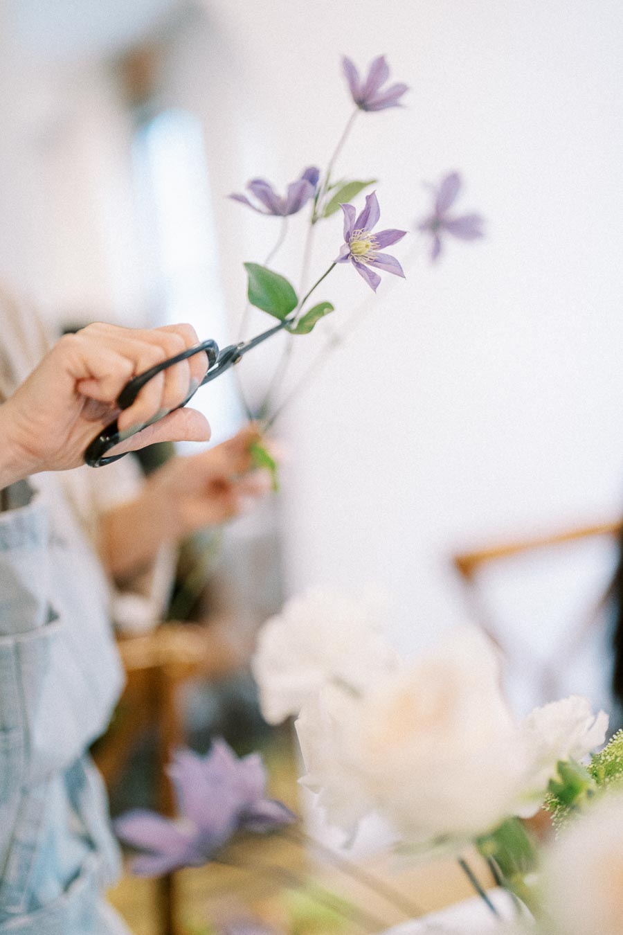 A person trimming purple clematis flowers with scissors in a bright room, focusing on floral arrangement and delicate craftsmanship.