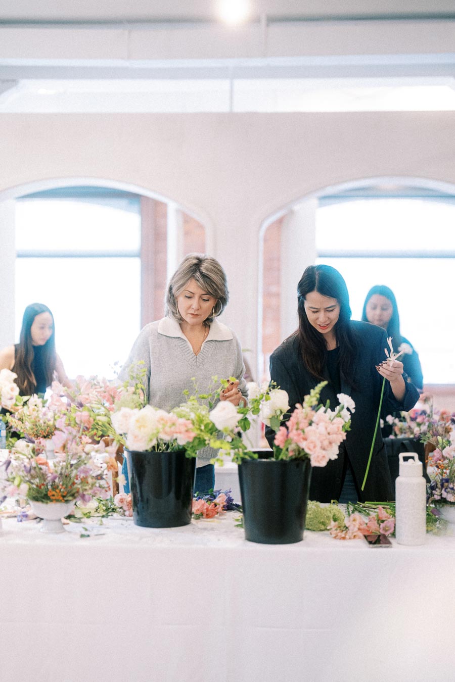 Two women participating in a floral arrangement workshop, arranging colorful flowers on a table in a bright, spacious room with large windows.