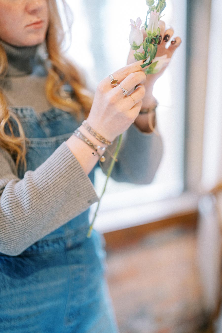 Young woman in a grey sweater and denim overalls holding a delicate flower indoors, showcasing bohemian jewelry.