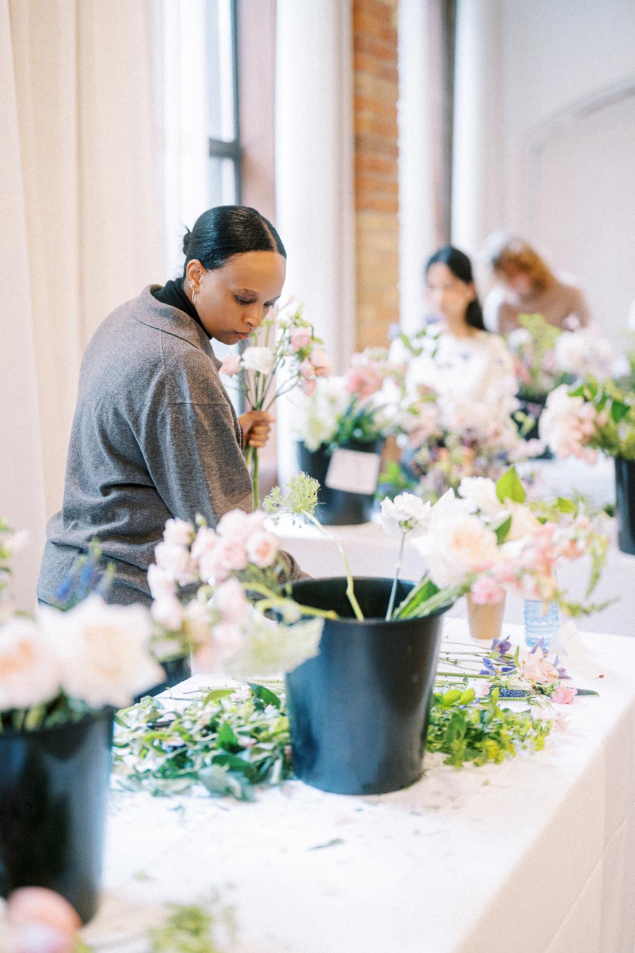 Florist arranging pink and white flowers in a bucket during a workshop with blurred participants and floral supplies in the background.