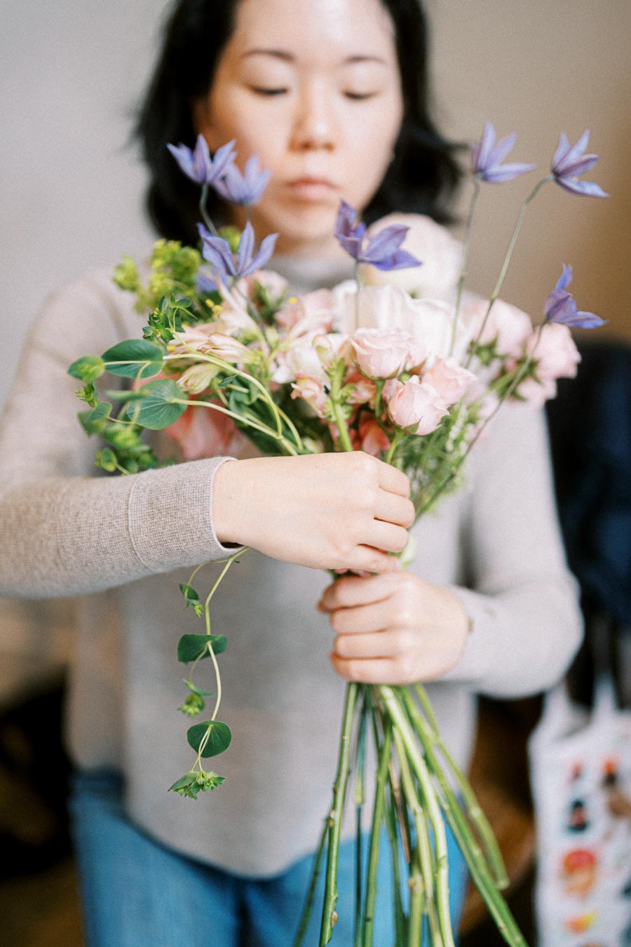A person arranging a vibrant bouquet of flowers with purple and pink blooms, wearing a gray sweater, demonstrating floral design skills.