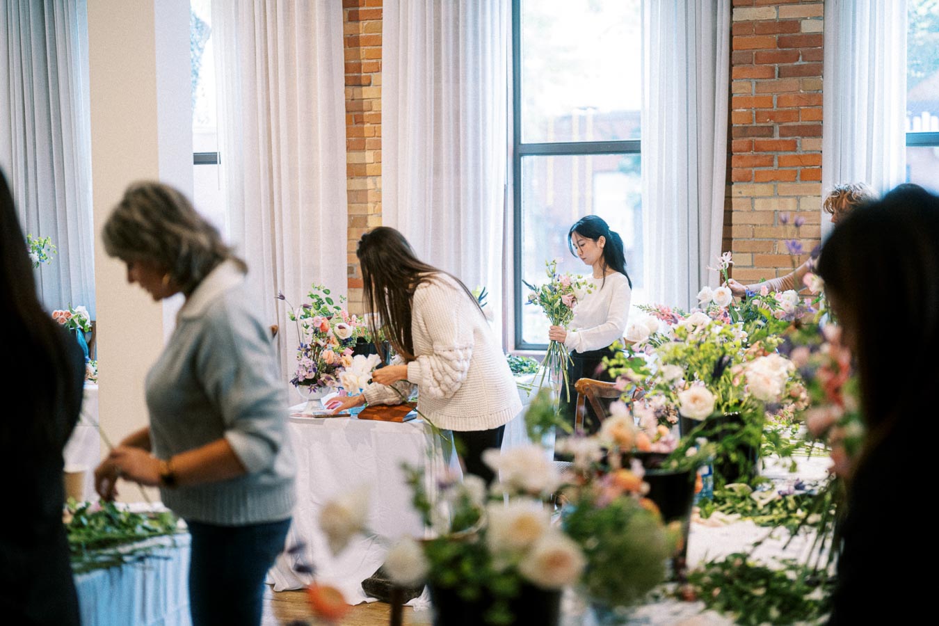 People arranging flowers in a bright room with tables covered in floral arrangements and supplies, emphasizing a creative workshop or floral design class atmosphere.