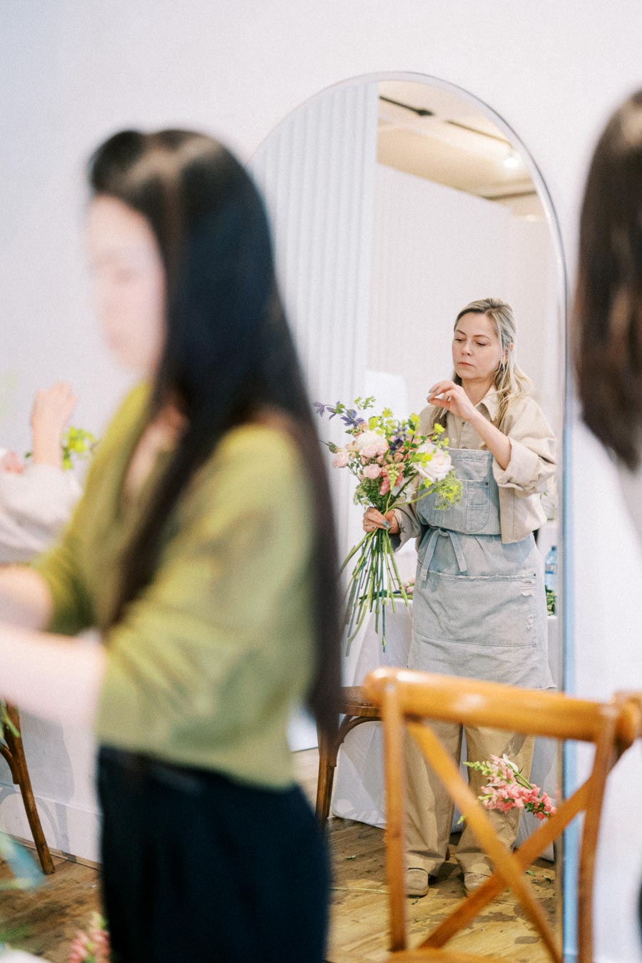 A woman wearing an apron arranges a bouquet of colorful flowers in a bright room with a mirror reflecting her actions, while another blurred figure works in the foreground, suggesting a collaborative floral design workshop.