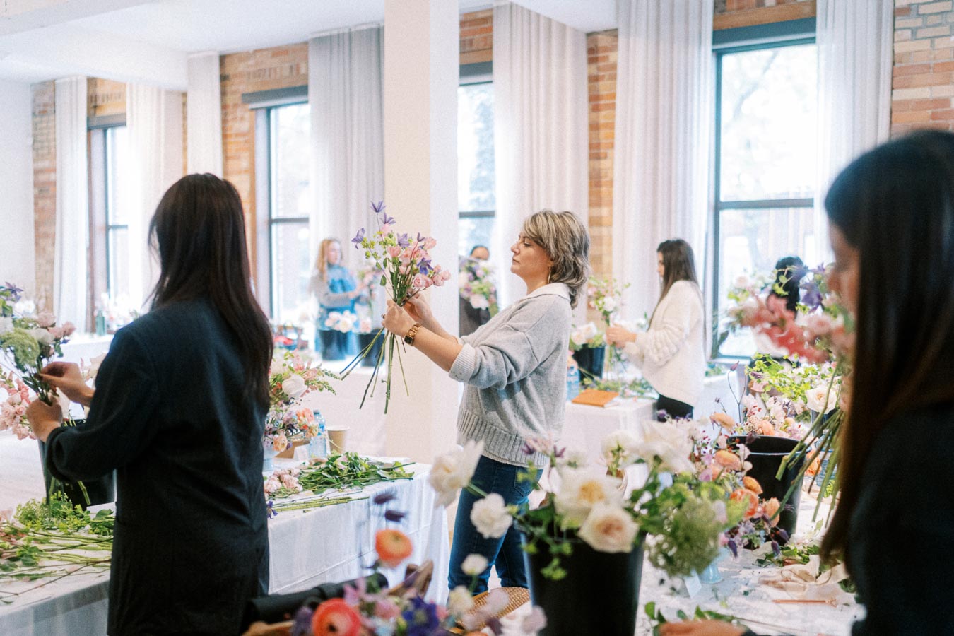 Women arranging colorful flowers in a workshop setting with natural light and wooden interiors.