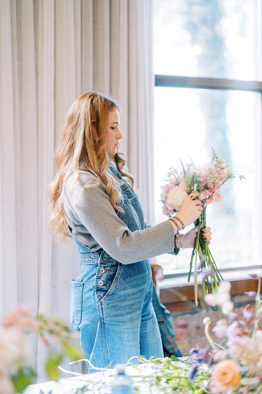 Woman arranging a bouquet of pink and white flowers indoors, wearing denim overalls and a gray sweater, with soft natural light from a large window.