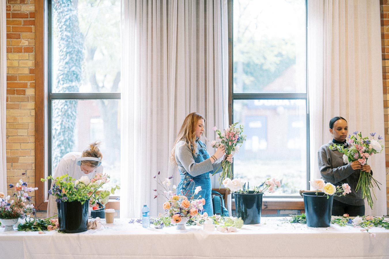 Three people arranging floral bouquets at a workshop, with vibrant flowers and tools spread across a table in a bright room with large windows.