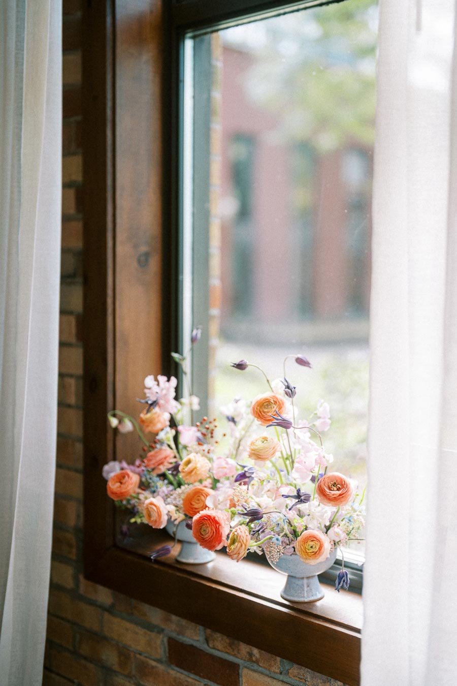 Elegant floral arrangement with orange and pink flowers in blue vases on a windowsill, surrounded by sheer white curtains, and a view of greenery through the window.