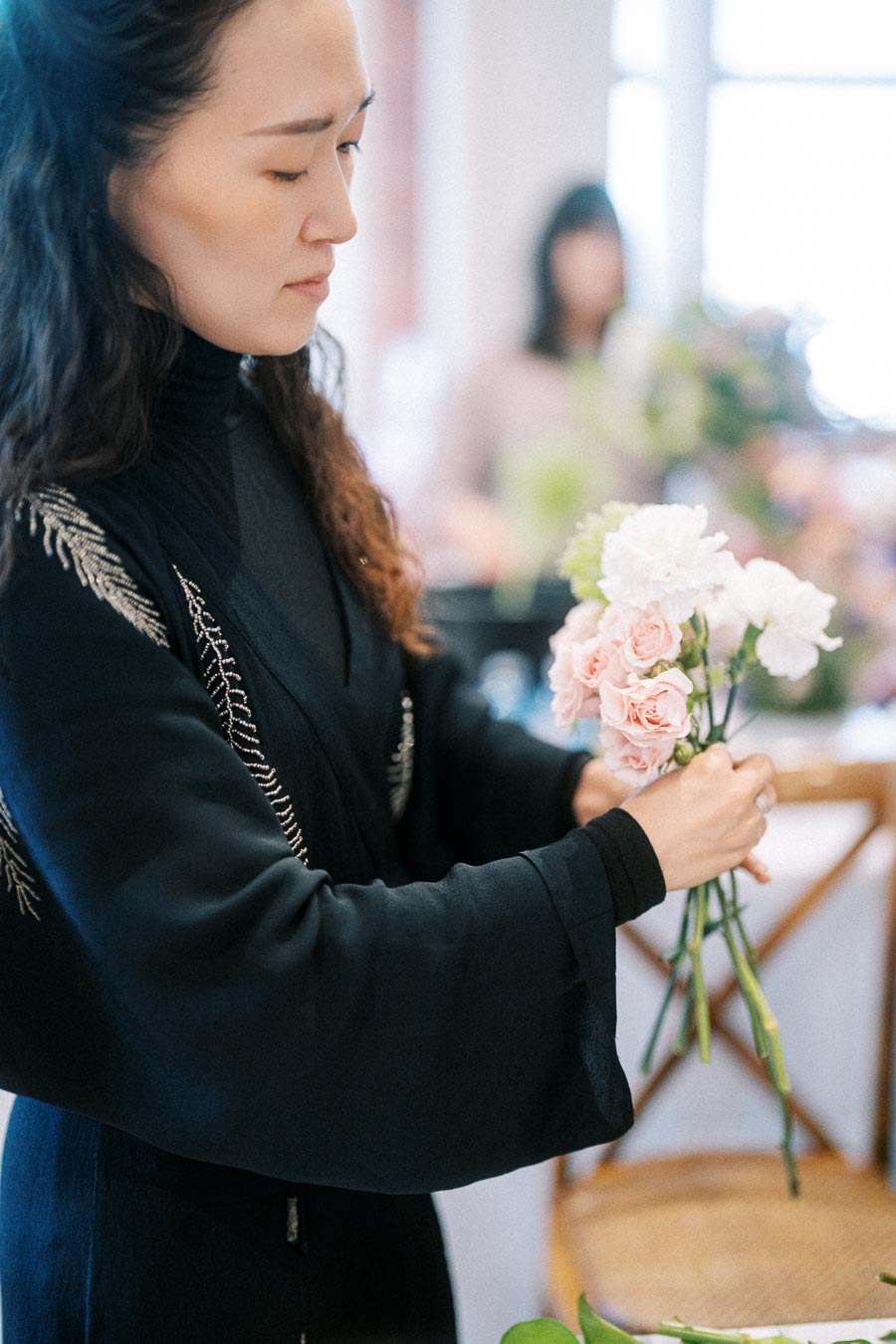 Florist arranging a bouquet of pink and white flowers in a bright room, wearing a black embroidered kimono.