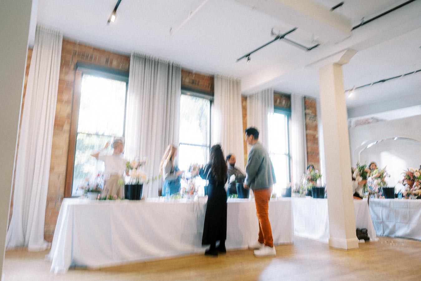 Blurry image of people socializing at a flower arrangement event in a bright, modern room with tall windows and white curtains.