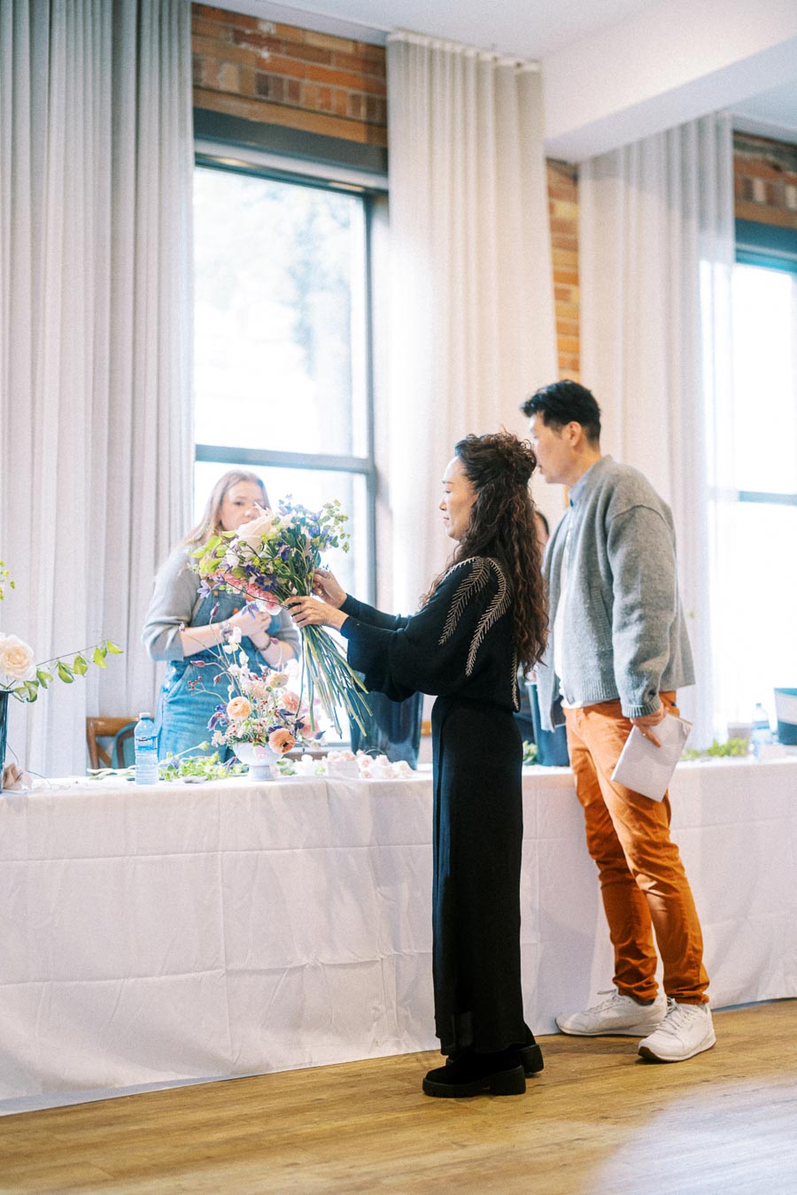 Florist arranging a bouquet at a floral workshop with participants observing, bright indoor setting with natural light from large windows.