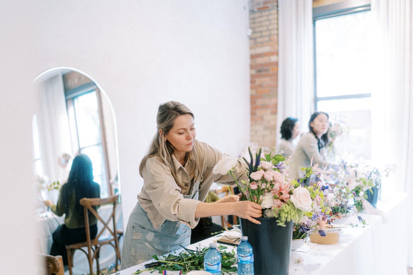 A woman arranging a bouquet of flowers in a bright, modern workshop setting with a brick wall and large window in the background. She is focused on creating an elegant floral display, surrounded by various flowers and greenery on the table.