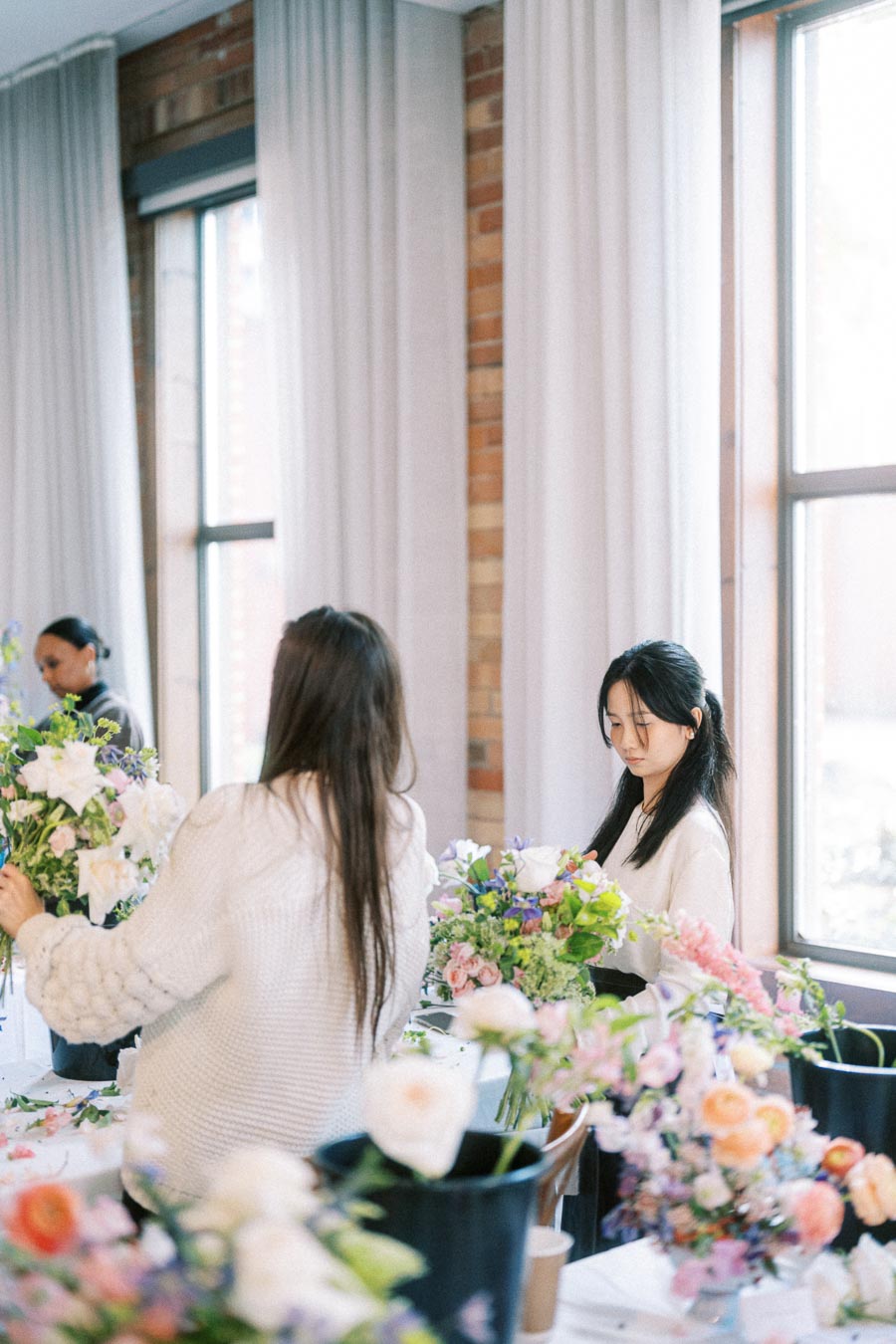 Two women arrange colorful floral bouquets in a bright room with large windows, showcasing a creative flower workshop setting.