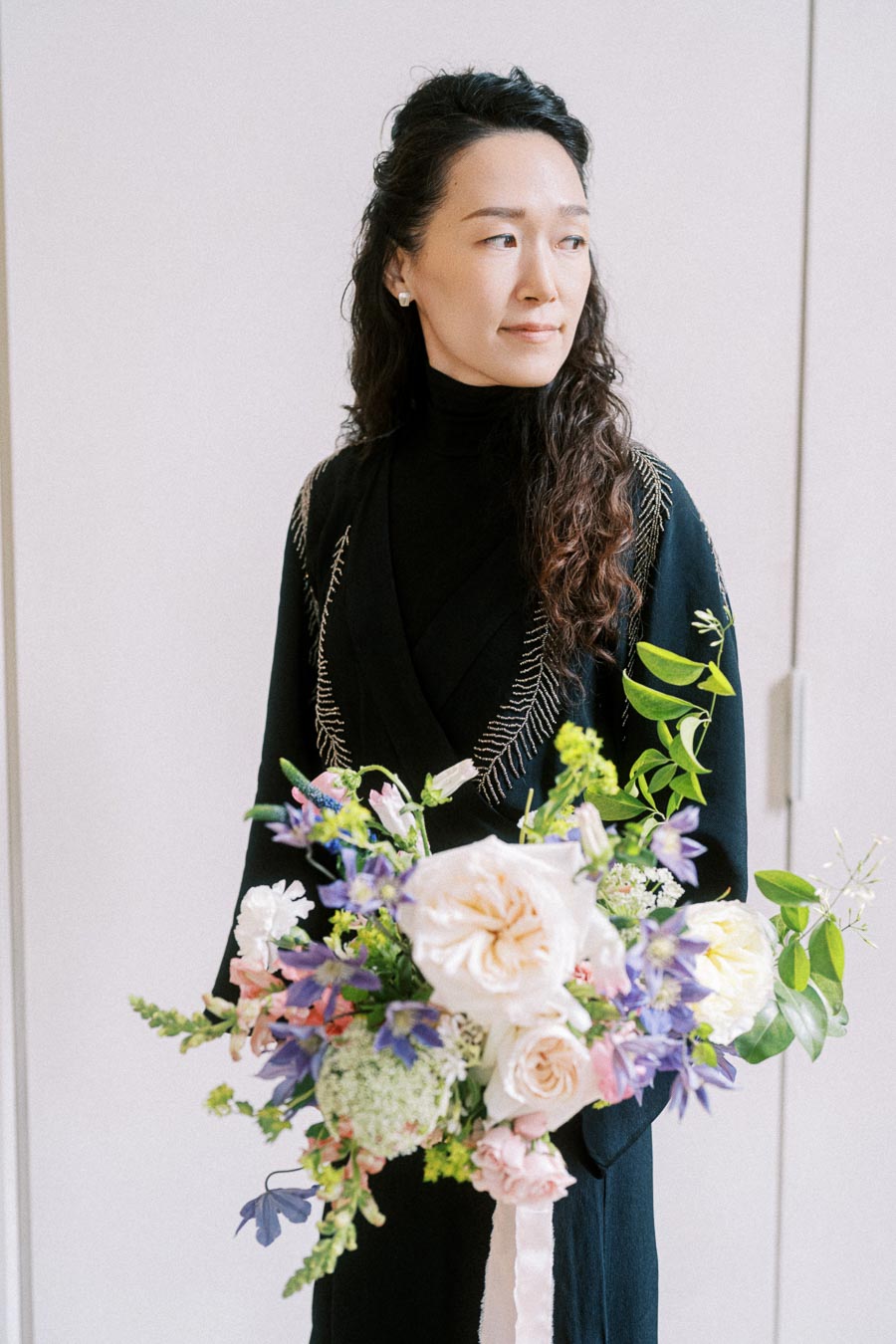 A woman in a black dress holding a colorful bouquet of flowers, featuring white, pink, and purple blooms, with green leaves, standing against a light background.