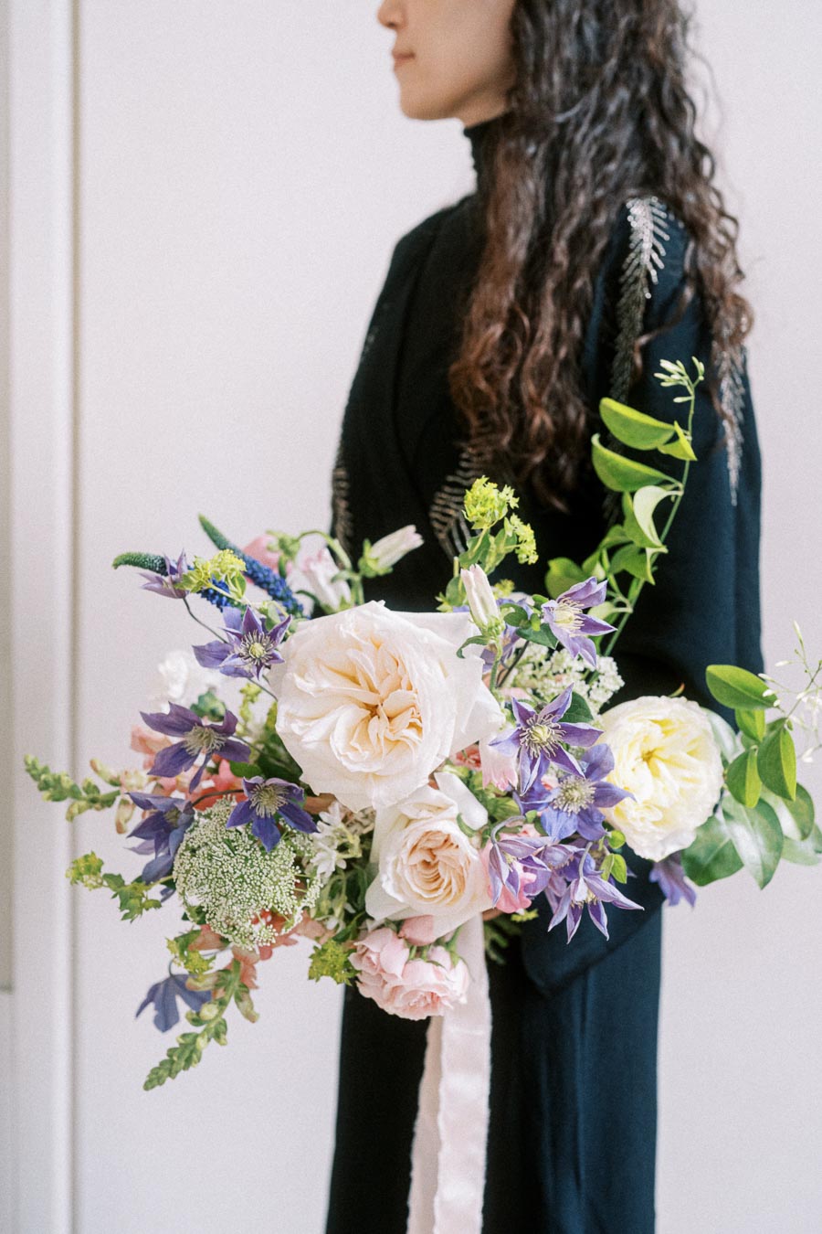 A woman in a black dress holding a vibrant bouquet of flowers, featuring white and pink roses with purple accents and lush greenery, against a neutral background.