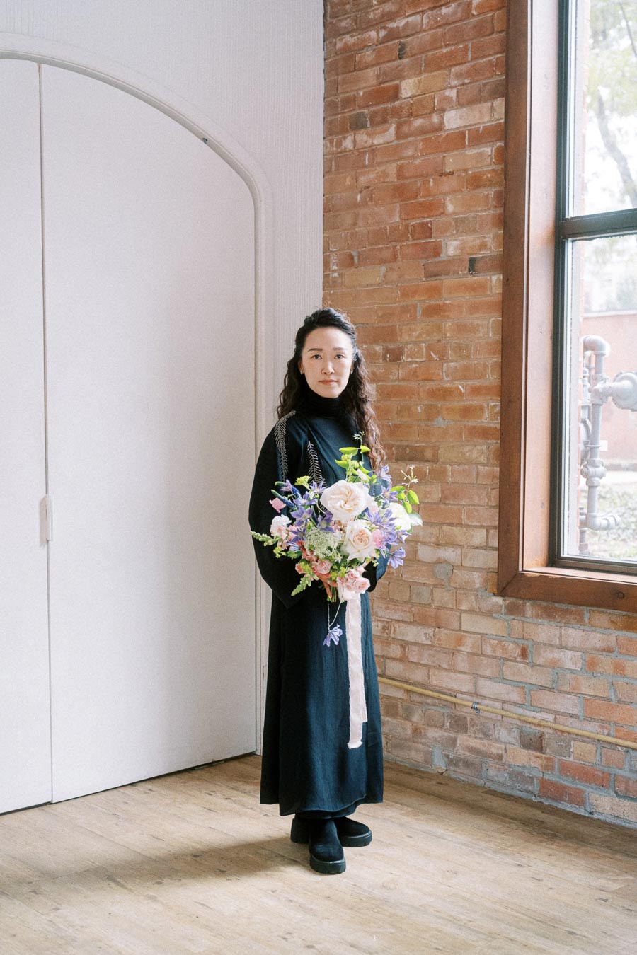 A woman in a black dress holds a colorful bouquet of flowers, standing in an industrial-style room with a brick wall and a large window.