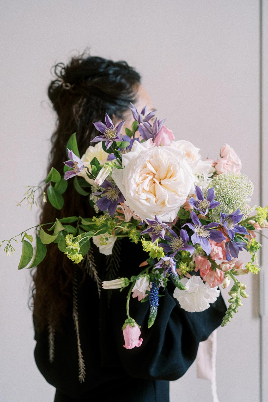 Floral bouquet with white and pastel pink roses, purple clematis, and assorted greenery held by a woman in a black dress, set against a neutral background.