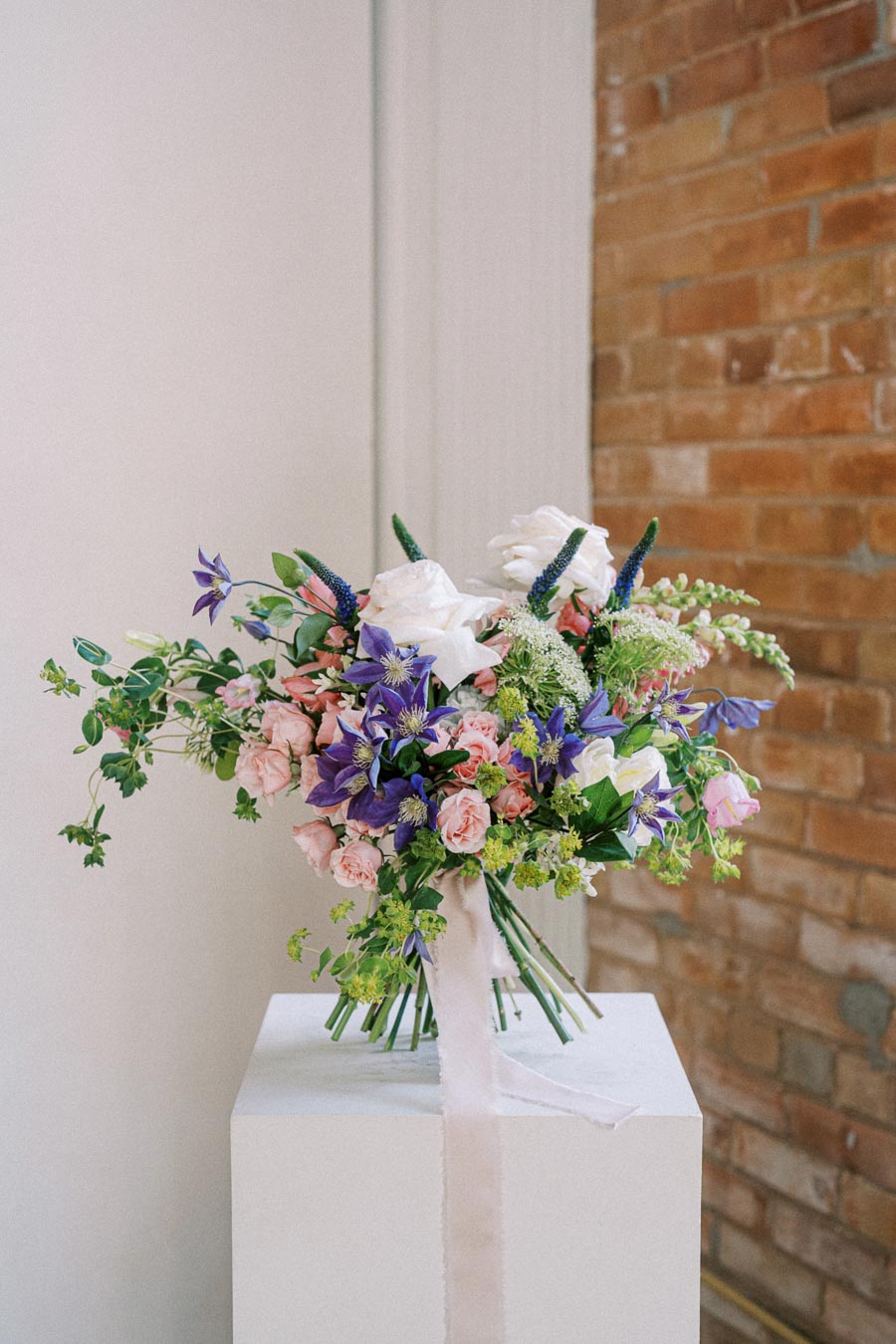 A vibrant flower bouquet with mixed blooms including pink roses, purple clematis, and white flowers, elegantly arranged with green foliage, set against a rustic brick wall and white pedestal.