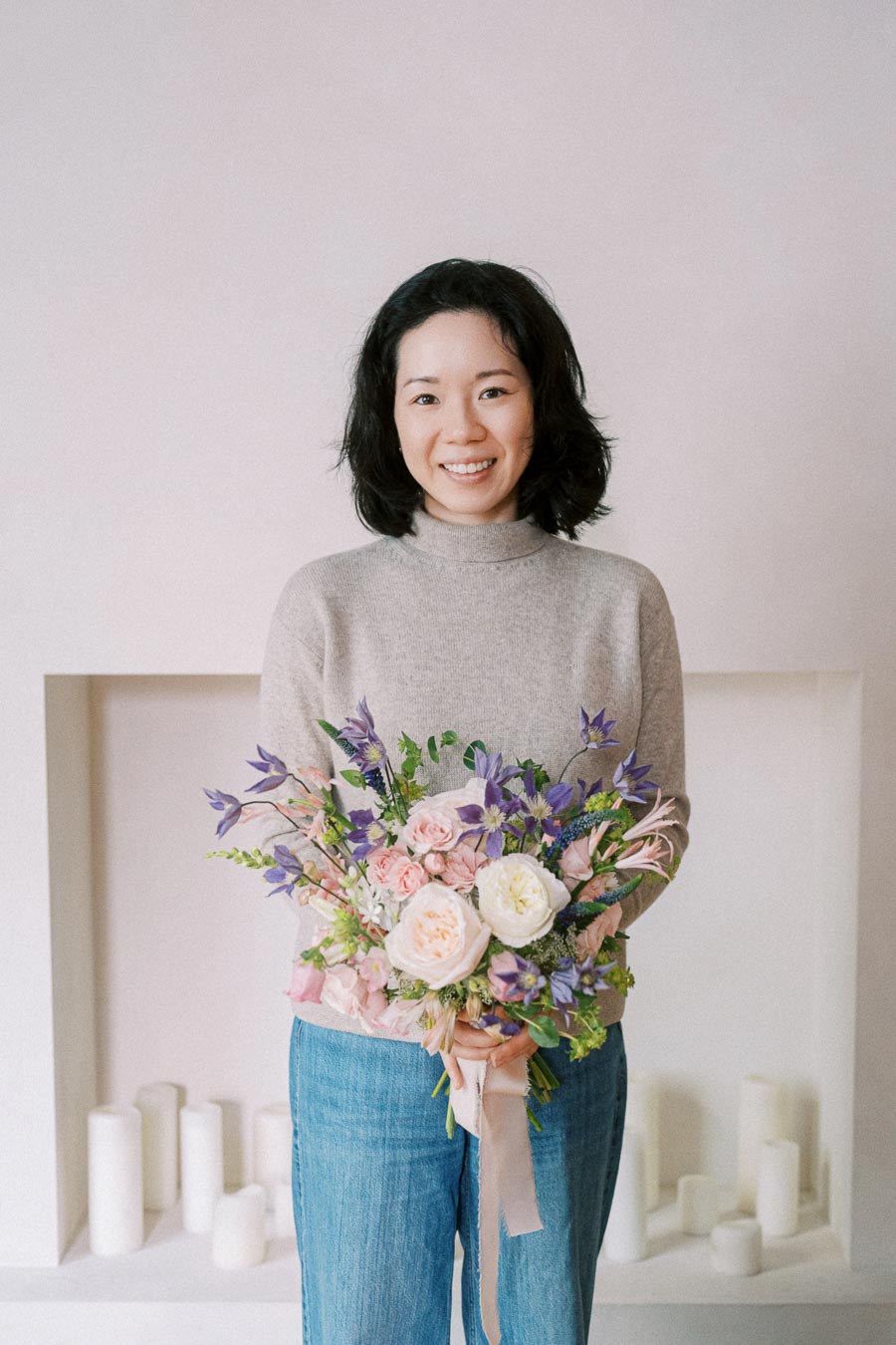 Smiling person holding a bouquet of pastel flowers, including roses and greenery, in a minimalistic room with white candles in the background.