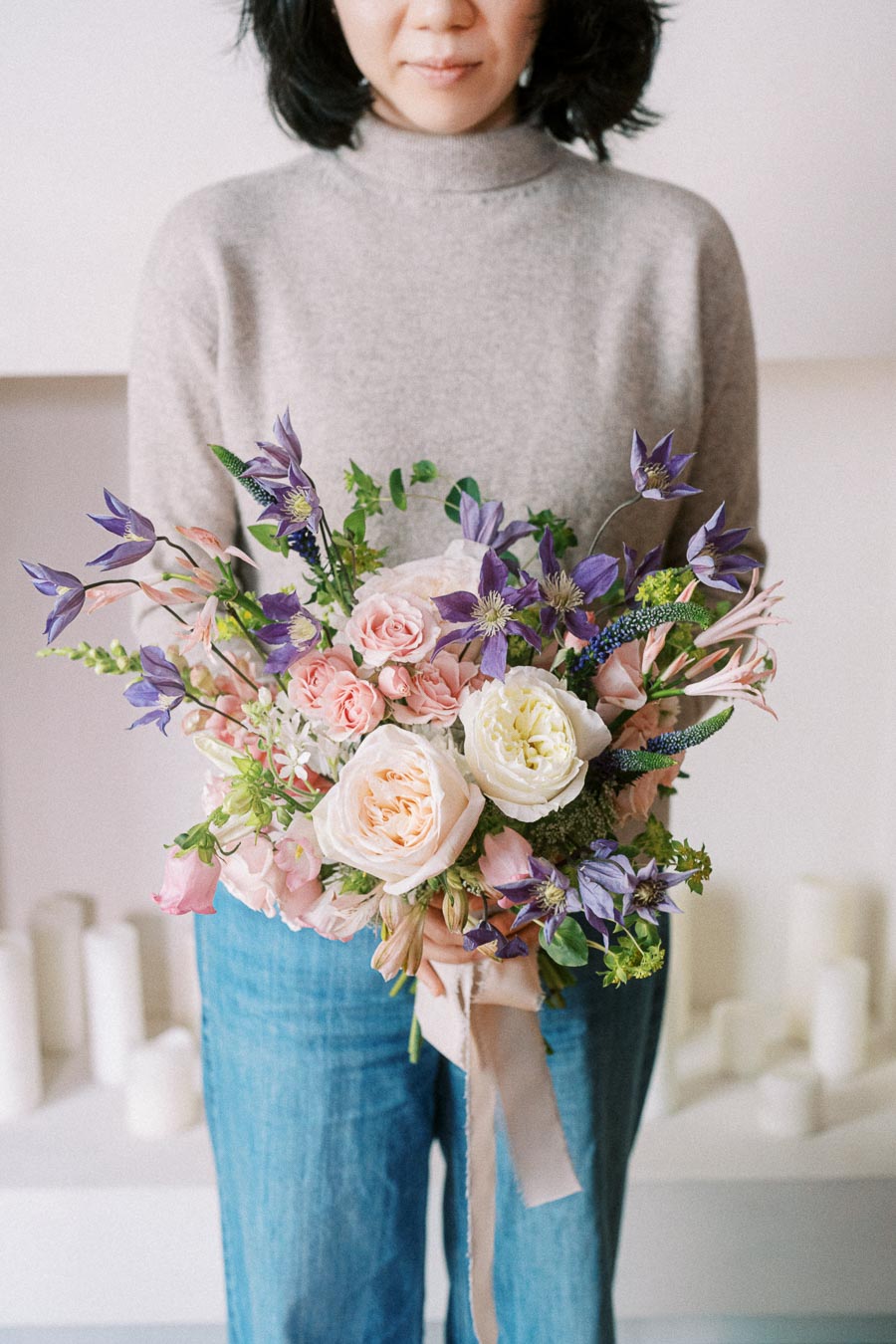 Person holding a colorful floral bouquet with pink, white, and purple flowers, wearing a gray sweater and jeans, in a bright indoor setting.