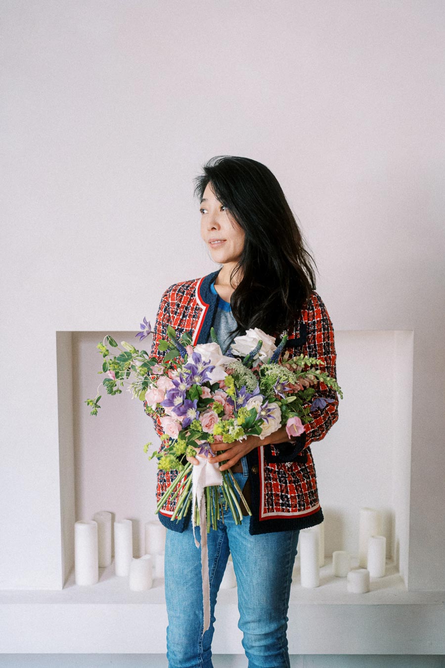 A person in a colorful jacket holding a vibrant bouquet of flowers, standing in front of a minimalist white background with white candles.