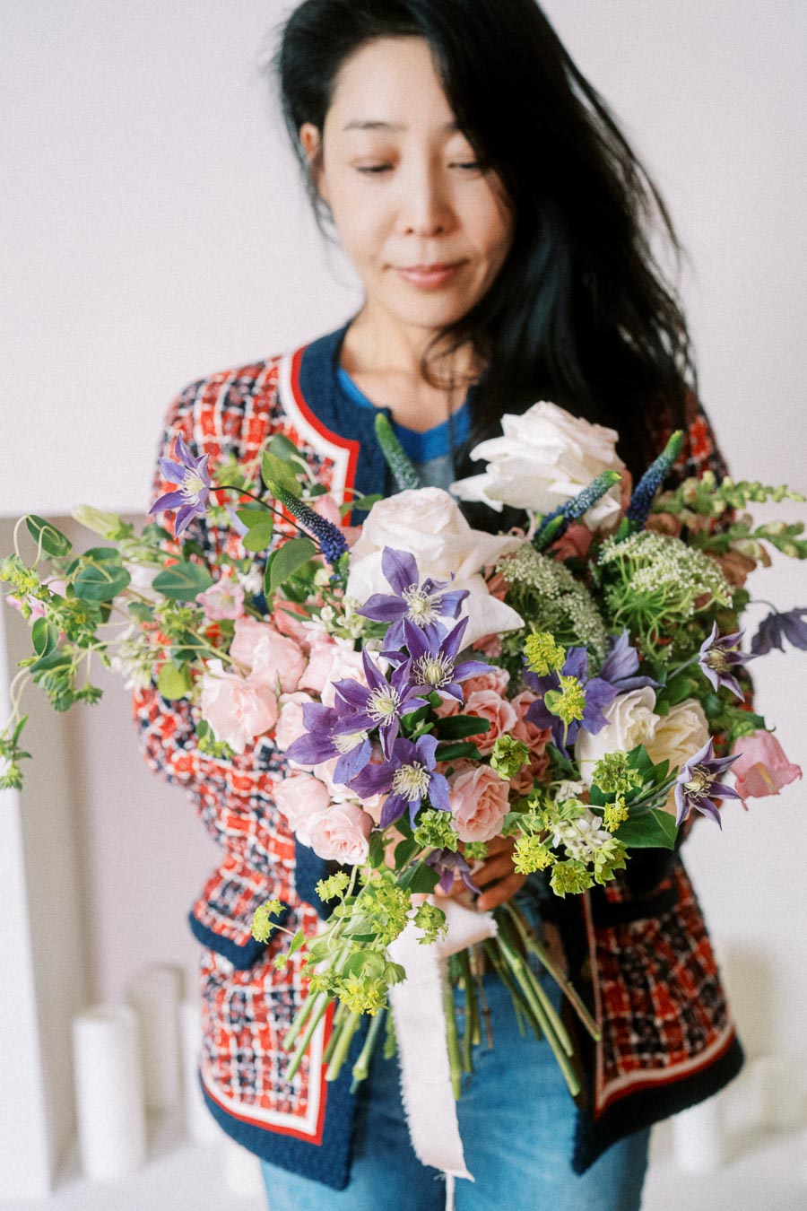 A person wearing a patterned jacket holding a vibrant bouquet of mixed flowers including purple clematis, white roses, and green foliage against a soft background.