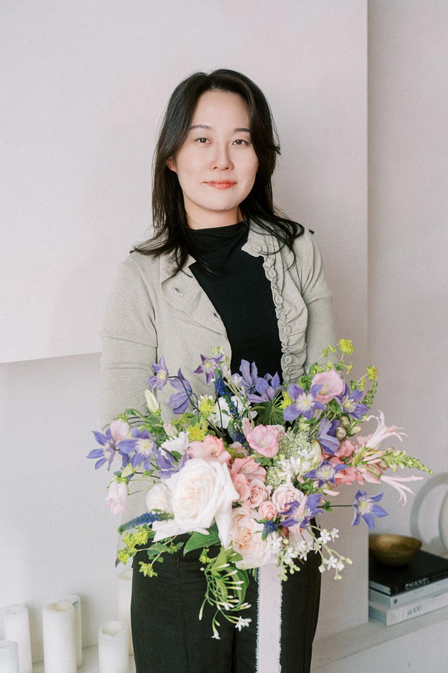 A person holding a vibrant bouquet of pastel flowers, including roses and lavender blooms, standing indoors beside a stack of books and decorative candles.