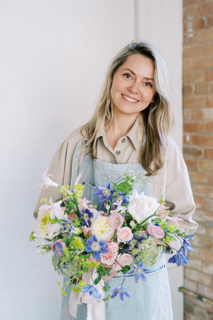 Woman smiling while holding a colorful floral bouquet, wearing a light blue apron; brick wall in the background.